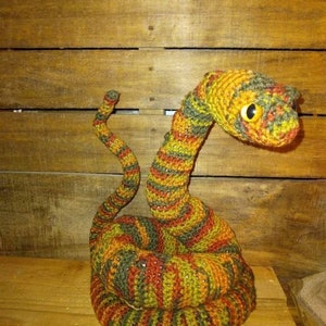May include: A crocheted snake toy with a striped pattern in shades of green, orange, and brown. The snake is curled up in a coil with its head raised, and it has a large, round, yellow eye.