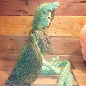 May include: A green crocheted stuffed toy with a long tail and a hooded cloak. The toy is sitting on a wooden surface.