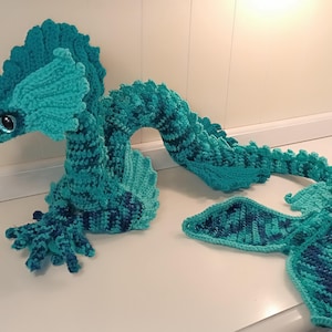 May include: A handmade crochet seahorse in shades of teal and blue. The seahorse has a detailed head, a curved body, and a fan-shaped tail. The plush toy is displayed on a white surface.