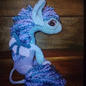 May include: A crocheted plush toy in the shape of a mythical creature with blue, purple, and white yarn. The creature has a long tail, large ears, and a single eye.