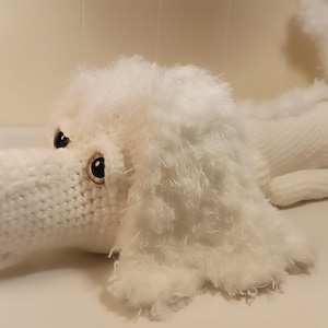 May include: A white crocheted stuffed dog with black eyes and a red tongue. The dog is lying on its side with its head turned to the left.
