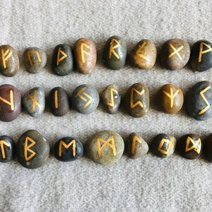 Może przedstawiać: Zestaw 24 gładkich, szarych kamieni z namalowanymi złotymi runami. Każdy kamień przedstawia inny symbol runy.