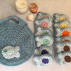 May include: A blue crocheted bag with a fish design, alongside a set of matching coasters with colourful centres. A candle and seashells are also visible. The items are handmade.