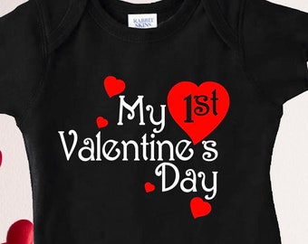 May include: Black baby onesie with white text that reads "My 1st Valentine's Day" with a red heart around the "1st". There are several small red hearts scattered around the text.