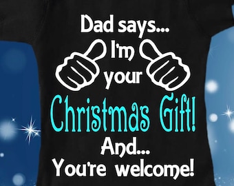 Puede incluir: Un body negro para bebé con texto blanco que dice "Papá dice... ¡Soy tu regalo de Navidad! Y... ¡Bienvenido!" El texto está rodeado de dos pulgares hacia arriba blancos.
