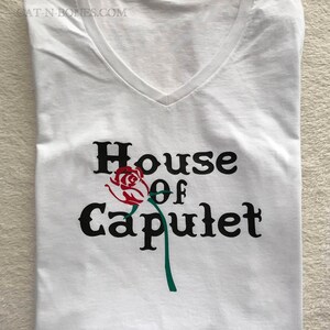 May include: White v-neck t-shirt with a black graphic that says "House of Capulet" with a red rose and green stem.