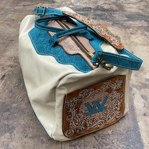 Op de afbeelding: Een witte leren reistas met een blauw en bruin bloemenpatroon. De tas heeft een ritssluiting en een bovenste handvat. De tas heeft een grote, bruine leren patch met een gestileerd "W"-logo.