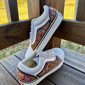 以下が含まれることがあります： グレーのスニーカーのペアで、カスタムレザーのディテールが施されています。デザインは、茶色、ピンク、緑の色合いのフローラルとハートのパターンが特徴です。靴は、黒いストライプとグレーの靴ひもが付いた白いソールです。