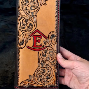 May include: A tan leather long wallet featuring intricate hand-tooled floral scrollwork patterns. The center displays a bold, red-painted letter E enclosed within a red geometric frame. The edges are stitched with vibrant red thread.