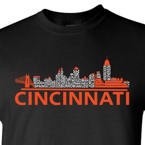 May include: Black t-shirt featuring an orange silhouette of the Cincinnati skyline. The skyline is made up of words associated with the city, including "Burrow", "Bengals", and "Reds". The text "CINCINNATI" is printed below the skyline.