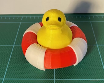 May include: A yellow rubber duck toy sits inside a red, white, and red life preserver. The duck has black eyes and a small beak. The life preserver is round and has three bands of color. The toy is on a green surface.