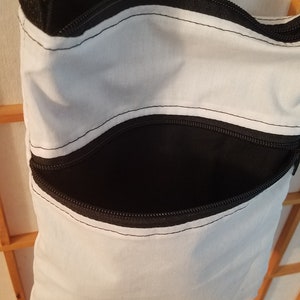 May include: A white canvas crossbody bag with two black zippered pockets. The bag has a black adjustable strap.