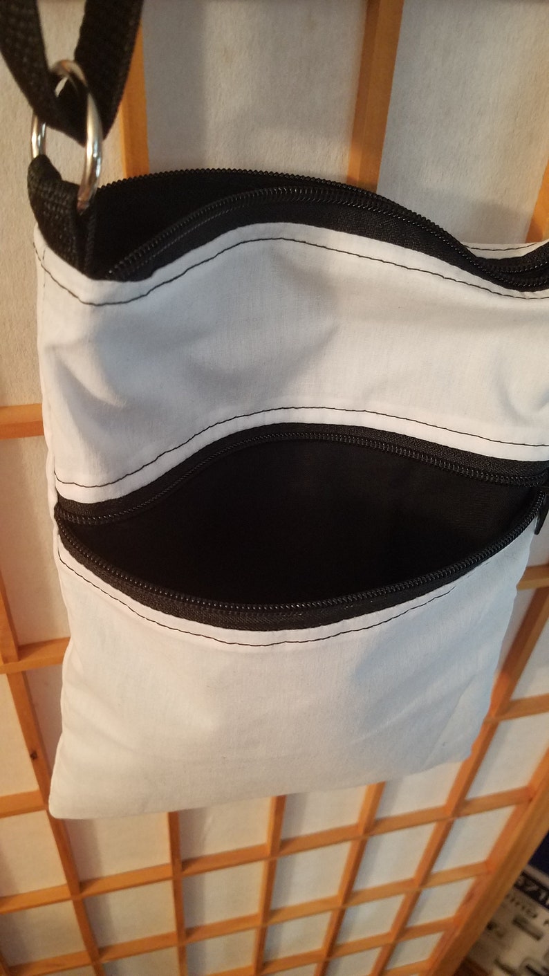 May include: A white canvas crossbody bag with two black zippered pockets. The bag has a black adjustable strap.