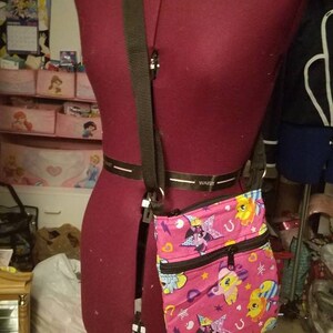 May include: A pink crossbody bag with a black strap. The bag has a colorful print of cartoon horses with stars and hearts. The bag has two zippered compartments.