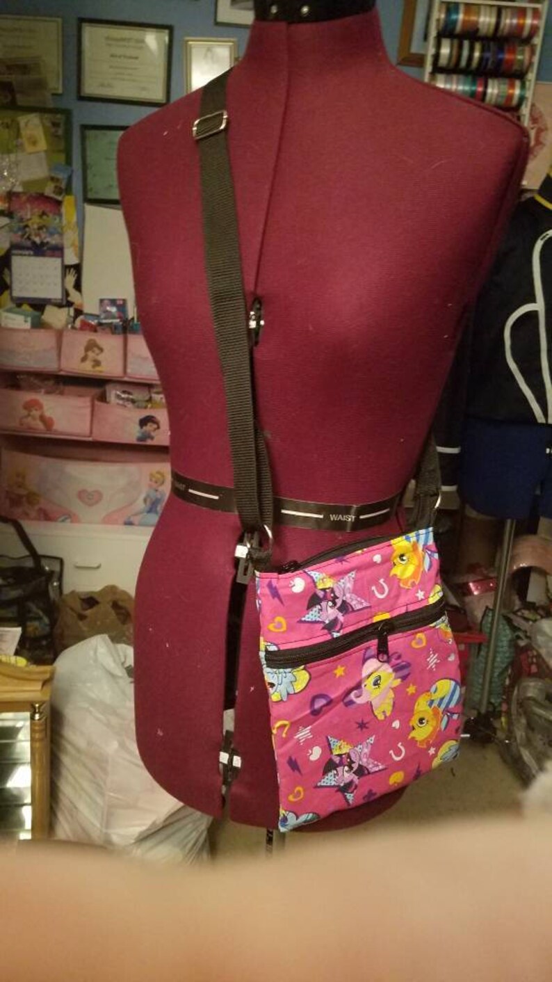 May include: A pink crossbody bag with a black strap. The bag has a colorful print of cartoon horses with stars and hearts. The bag has two zippered compartments.