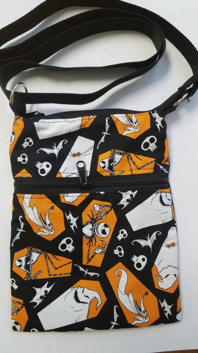 May include: Black and orange fabric crossbody bag with a zipper closure. The bag features a repeating pattern of Jack Skellington and other characters from The Nightmare Before Christmas.