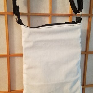May include: A white canvas crossbody bag with a black zipper and black adjustable strap. The bag is hanging on a wooden slat wall.