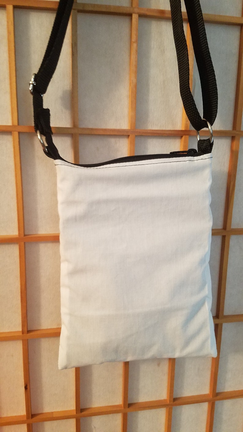 May include: A white canvas crossbody bag with a black zipper and black adjustable strap. The bag is hanging on a wooden slat wall.