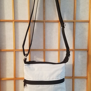 May include: A white crossbody bag with black straps and two zippered compartments. The bag is hanging on a wooden screen.
