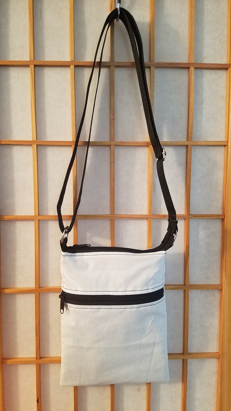 May include: A white crossbody bag with black straps and two zippered compartments. The bag is hanging on a wooden screen.