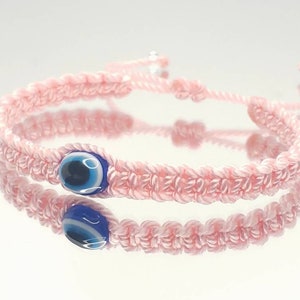 May include: A pink string bracelet with a blue evil eye charm. The bracelet is made of a braided string and has a simple design.