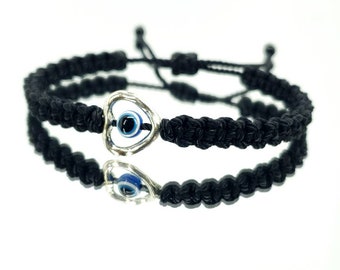 May include: Black macrame bracelet with a silver heart-shaped charm featuring a blue and white evil eye. The bracelet is adjustable and sits on a reflective surface.