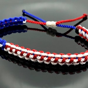 K&ouml;nnte beinhalten: Ein geflochtenes Freundschaftsarmband in Rot, Wei&szlig; und Blau. Das Armband ist mit einer Knotentechnik hergestellt, die ein gewebtes Muster erzeugt.