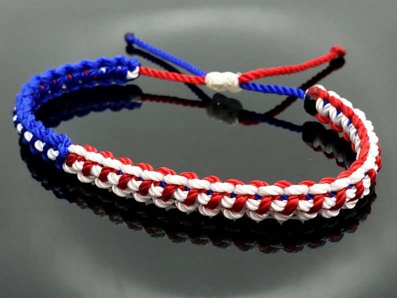 K&ouml;nnte beinhalten: Ein geflochtenes Freundschaftsarmband in Rot, Wei&szlig; und Blau. Das Armband ist mit einer Knotentechnik hergestellt, die ein gewebtes Muster erzeugt.