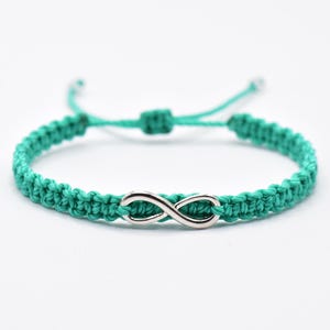 May include: A teal braided cord bracelet featuring a central silver-toned infinity symbol charm. The bracelet is constructed with a woven knot pattern and includes adjustable pull strings with small knots at the ends for a secure fit.