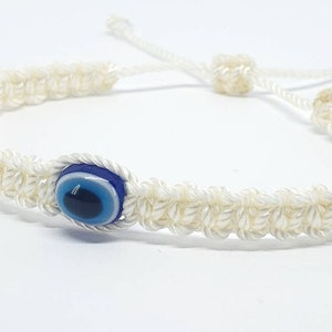 May include: Cream-colored macrame bracelet featuring a blue and white evil eye charm. The bracelet is adjustable with a sliding knot closure. The woven design is visible, and the bracelet is displayed on a white background.