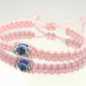 May include: A pink macrame bracelet with a blue and white bead, accented with silver-colored rhinestones. The bracelet is adjustable with a sliding knot closure. The bracelet is displayed on a reflective surface.