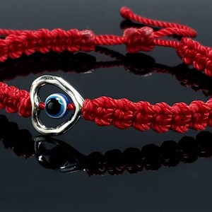K&ouml;nnte beinhalten: Rotes Armband mit einem silbernen Herz-Anh&auml;nger mit einem blauen b&ouml;sen Auge. Das Armband ist ein beliebtes Symbol f&uuml;r Schutz und Gl&uuml;ck.