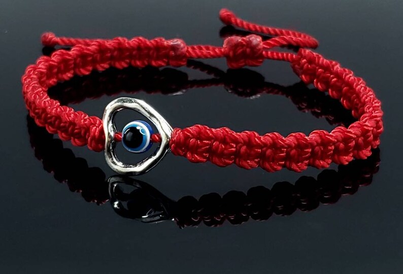 K&ouml;nnte beinhalten: Rotes Armband mit einem silbernen Herz-Anh&auml;nger mit einem blauen b&ouml;sen Auge. Das Armband ist ein beliebtes Symbol f&uuml;r Schutz und Gl&uuml;ck.
