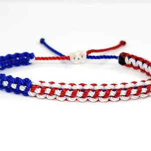 K&ouml;nnte beinhalten: Ein geflochtenes Freundschaftsarmband in Rot, Wei&szlig; und Blau. Das Armband ist mit einem gewebten Muster gefertigt und hat einen einfachen Knotenverschluss.