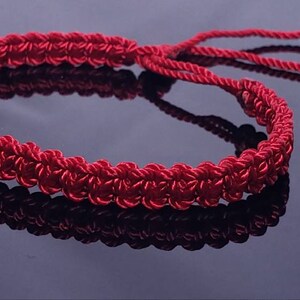 Puede incluir: Un brazalete trenzado rojo con dijes de coraz&oacute;n plateados. El brazalete est&aacute; atado con un nudo y tiene una borla.