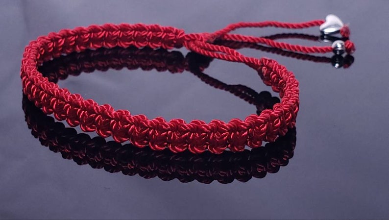 Puede incluir: Un brazalete trenzado rojo con dijes de coraz&oacute;n plateados. El brazalete est&aacute; atado con un nudo y tiene una borla.