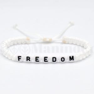 May include: White macrame bracelet with the word "FREEDOM" spelled out in black letters on white beads. The bracelet is adjustable and sits against a white background.