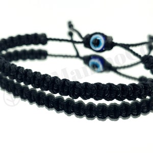 May include: Two black braided bracelets with blue evil eye charms. The bracelets are tied together with a knot.