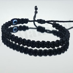 May include: Two black braided bracelets with blue evil eye charms. The bracelets are tied together and are on a white surface.