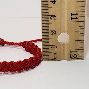 Puede incluir: Un brazalete de cuerda roja con cierre de nudo, medido con una regla. El brazalete tiene un di&aacute;metro de aproximadamente 2,5 cm.