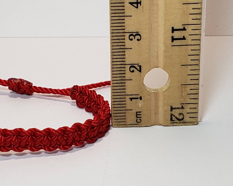 Puede incluir: Un brazalete de cuerda roja con cierre de nudo, medido con una regla. El brazalete tiene un di&aacute;metro de aproximadamente 2,5 cm.