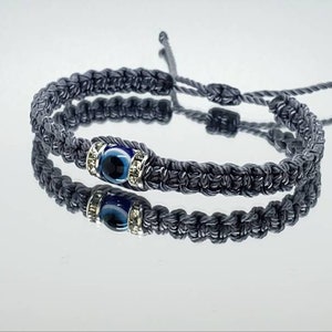 May include: Two gray woven bracelets with adjustable ties. Each bracelet features a central blue eye charm with silver-colored accents. The bracelets are displayed on a reflective surface, creating a mirrored effect.