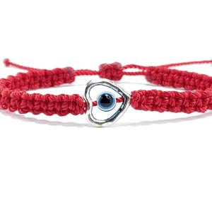 K&ouml;nnte beinhalten: Rotes Armband mit einem silbernen Herz-Anh&auml;nger mit einem blauen b&ouml;sen Auge. Das Armband ist gekn&uuml;pft und hat einen verschiebbaren Knoten zum Anpassen der Gr&ouml;&szlig;e.