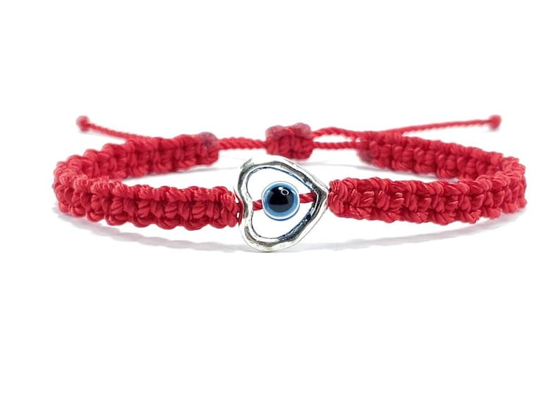 K&ouml;nnte beinhalten: Rotes Armband mit einem silbernen Herz-Anh&auml;nger mit einem blauen b&ouml;sen Auge. Das Armband ist gekn&uuml;pft und hat einen verschiebbaren Knoten zum Anpassen der Gr&ouml;&szlig;e.
