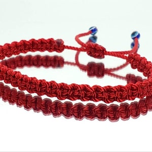 May include: Red string bracelet with two blue evil eye beads. The bracelet is knotted and has a sliding knot for adjusting the size.
