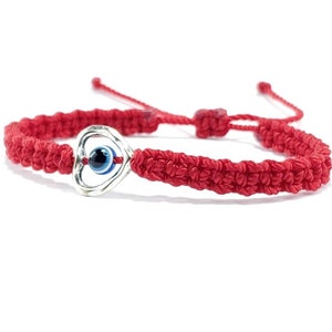 K&ouml;nnte beinhalten: Rotes Armband mit einem silbernen Herz-Anh&auml;nger mit einem blauen b&ouml;sen Auge. Das Armband ist mit einem Knoten gebunden und hat einen verschiebbaren Knoten zum Anpassen der Gr&ouml;&szlig;e.