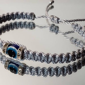 May include: A gray braided bracelet with a blue evil eye charm. The charm is surrounded by clear rhinestones.