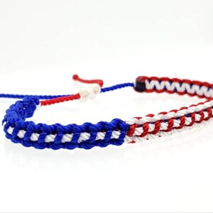 K&ouml;nnte beinhalten: Ein geflochtenes Freundschaftsarmband in Blau, Wei&szlig; und Rot. Das Armband besteht aus drei Schn&uuml;ren, die zu einem Muster geflochten sind, das an eine Flagge erinnert.