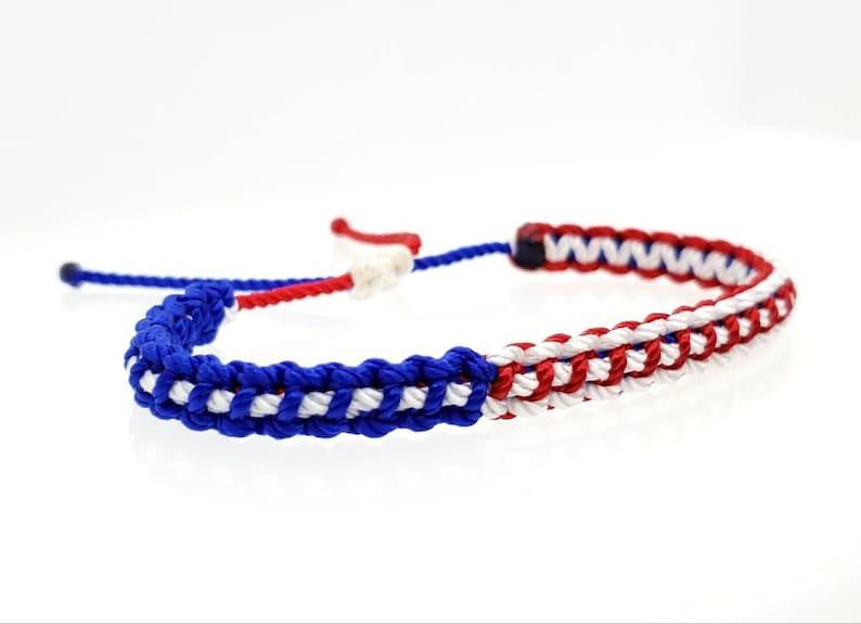K&ouml;nnte beinhalten: Ein geflochtenes Freundschaftsarmband in Blau, Wei&szlig; und Rot. Das Armband besteht aus drei Schn&uuml;ren, die zu einem Muster geflochten sind, das an eine Flagge erinnert.