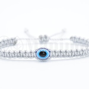 May include: A gray braided bracelet with a blue evil eye charm. The bracelet is adjustable with a sliding knot.